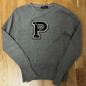 Stickad Ralph Lauren Tröja - Grå stickad tröja från Polo Ralph Lauren med svartvit P-applikation. Klassisk college-stil med rund hals och ribbade muddar. Midjan har krympt något – perfekt för slim fit och passar även Small.