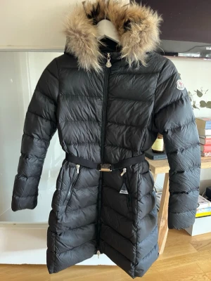 Svart lång barn dunjacka från Moncler - Snygg svart lång dunjacka från Moncler med bälte i midjan och luva med fluffig fuskpäls. Jackan är i BARNSTORLEK (12 år/152cm) Jackan har två dragkedjefickor framtill och Moncler-logga på ärmen. Jackan är köpt för ca 10 år sedan men är fortfarande i gott skick. Defekter som förekommer är följande: Repig/slitage på äkthets brickan på ena dragkedjan, Repor på metallen på bältet, Lös tråd på ärmarna. Om du vill ha fler bilder på defekter eller annat är det bara att skiva ett meddelande☺️ 