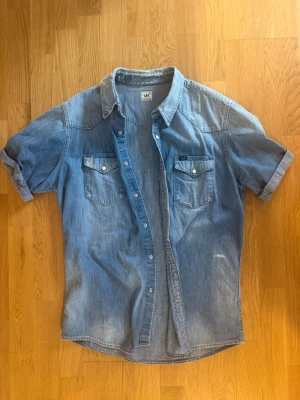 Blå jeansskjorta från Lee - Säljer en klassisk blå jeansskjorta från Lee med korta ärmar och två bröstfickor med tryckknappar. Skjortan har en avslappnad passform och tydliga sömmar som ger en snygg vintagekänsla. Perfekt till jeans eller shorts för en cool look.