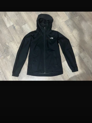 Svart regnjacka från The North Face - Snygg svart regnjacka från The North Face med huva och dragkedja framtill. Jackan har diskret logga på bröstet och är tillverkad i ett vattenavvisande material. Perfekt för regniga dagar och enkel att matcha med olika outfits. Har ett brännmärke från en platttång, vilket inte syns så mkt. Ett litet hål vid ärmen s går att sy om man skulle önska de. Nypris: runt 1700kr. Köptes i vintras. Funkar bra till hösten och vintern. 
