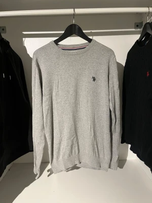 Grå stickad tröja U.S. Polo Assn. - Snygg grå stickad tröja från U.S. Polo Assn. med rund halsringning och diskret logga broderad på bröstet. Tröjan har långa ärmar och ribbade muddar vid ärmslut och nederkant. Perfekt till jeans eller chinos.