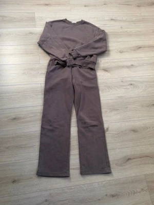Brunt loungewear-set med vida ben Gina tricot  - Mysigt loungewear-set i mjukt brunt tyg. Setet består av en långärmad sweatshirt med rund hals och ribbade muddar samt matchande byxor med vida ben. Perfekt för chill dagar hemma eller när du vill vara bekväm men ändå snygg.