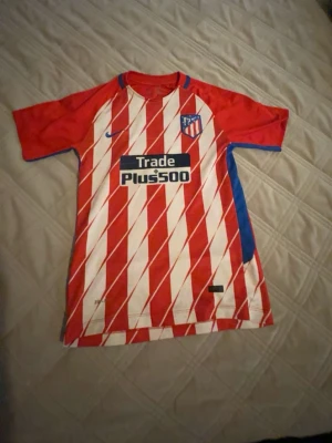 Atlético Madrid matchtröja Griezmann - Säljer en röd och vit Atlético Madrid matchtröja med Griezmann och nummer 7 på ryggen. Tröjan har sneda ränder, blå detaljer och klubbmärke på bröstet. Tillverkad i lätt och ventilerande material, perfekt för fotbollsträning eller match.