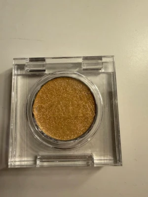 Guldig ögonskugga i singelförpackning - Snygg guldig ögonskugga från CAIA i en rund singelförpackning med transparent lock. Skuggan har en skimrig finish och är perfekt för att skapa en glittrig look. Förpackningen är stilren och enkel att ta med i väskan.