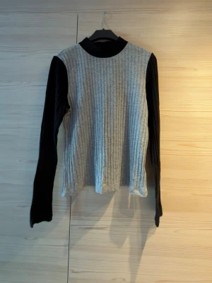 Grå ribbad tröja med svarta ärmar - Grå ribbad tröja med svarta långärmade ärmar. Tröjan har lite krage och är tillverkad i ett mjukt, stretchigt material. 