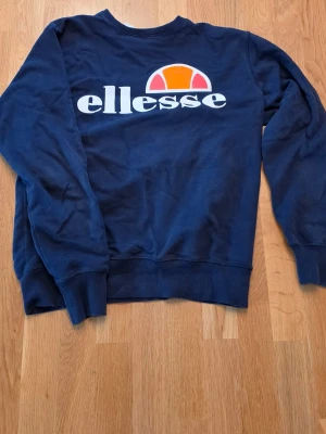 Mörkblå sweatshirt från Ellesse - Mörkblå sweatshirt från Ellesse med stort vitt och orange logotryck på bröstet. Tröjan har rund halsringning, ribbade muddar vid ärmslut och nederkant. 