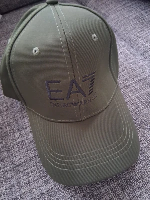 Olivgrön keps från EA7 Emporio Armani - Snygg olivgrön keps från EA7 Emporio Armani med broderad logga framtill. Klassisk böjd skärm och justerbart spänne bak. Tillverkad i slitstark bomull med ventilerande öljetter och detaljerad sömnad.