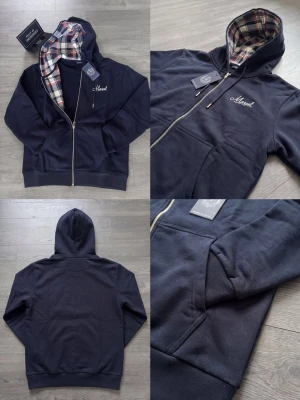 Mörkblå hoodie med rutigt foder - Stilren mörkblå hoodie med dragkedja och vit brodyr på bröstet. Insidan av huvan har ett snyggt rutigt foder i rött, vitt och svart. Modellen har fickor framtill och ribbade muddar vid ärmslut och nederkant. Perfekt för en avslappnad look.