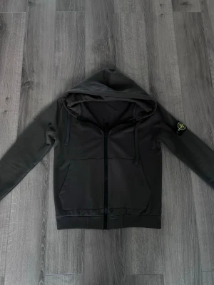 Stone island zip hoodie - Snygg svart hoodie med dragkedja framtill och stor huva. Hoodien har två rymliga fickor på magen och snörning i huvan. På vänster ärm sitter en patch med logga. Perfekt för en avslappnad och stilren look.