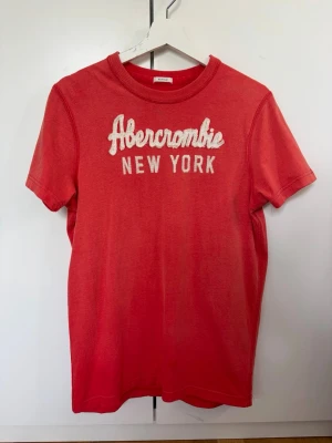 Röd Abercrombie & Fitch t-shirt - Snygg röd t-shirt från Abercrombie & Fitch med broderad text 'Abercrombie New York' på bröstet. Klassisk rund halsringning och korta ärmar. Perfekt till jeans eller shorts för en avslappnad look.