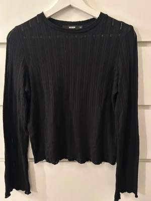 Svart ribbad långärmad topp Bik Bok - Svart långärmad topp från Bik Bok i ribbad, stretchig tyg. Enkel och stilren design med rund halsringning och lätt vågig kant vid ärmslut och nederkant och utsvängda armar. Perfekt att matcha med jeans eller kjol. Har en lite genomskinlig look. 