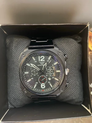  klocka Emporio Armani - Stilren svart klocka från Emporio Armani med fyrkantigt boett, svart länkarmband och flera små urtavlor. Klockan har tydliga vita siffror och markeringar samt silverfärgade detaljer på urtavlan. Perfekt accessoar för en modern look. Vet ej om det är äkta därför säljs den för billig.  DEN ÄR HELT NY