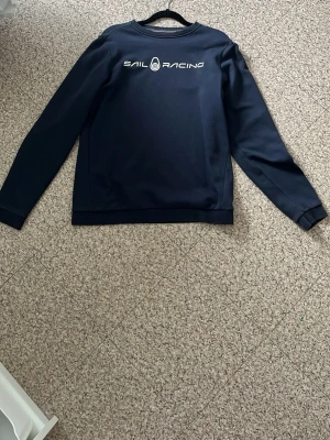 Marinblå Sail Racing sweatshirt - Snygg marinblå sweatshirt från Sail Racing med logga tryckt i vitt på bröstet och en mindre logga i nacken. Tröjan har rund hals, långa ärmar och ribbade muddar. Perfekt för en sportig och stilren look.