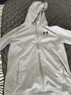 Ljusgrå vindjacka från Under Armour - Snygg ljusgrå vindjacka från Under Armour med huva och dragkedja framtill. Jackan har två fickor, logga på bröstet och stor text på ryggen. Perfekt för blåsiga dagar och sportiga outfits.