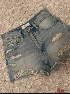 Blåa low waist jeansshorts från Zara - Säljer ett par ljusblåa, low jeansshorts från Zara med slitna detaljer och råa kanter. Shortsen har klassisk femficksdesign och dragkedja med knapp. Perfekta för sommaren och ger en avslappnad vibe.