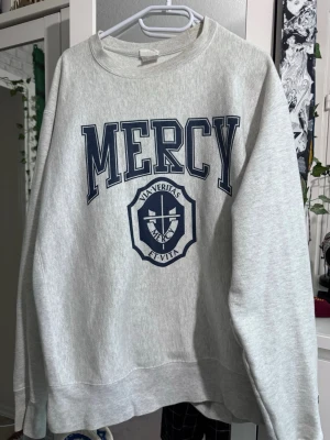 Grå sweatshirt Mercy från Champion - Säljer en grå sweatshirt från Champion med stor mörkblå MERCY-text och emblem på bröstet. Klassisk college-look med rund halsringning och ribbade muddar. Champion-logga broderad på ärmen. Perfekt för en chill och sportig stil.