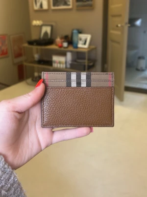 Brun kortfodral från Burberry - Säljer ett stilrent kortfodral från Burberry i brunt läder med klassiskt rutmönster upptill. Kortfodralet har flera fack för kort och är perfekt för dig som vill ha något både snyggt och praktiskt i fickan.