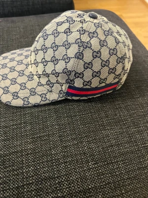 GG keps med blått GG-mönster - Säljer en snygg keps från GGi beige och blått med klassiskt GG-mönster över hela kepsen. På sidan finns en blå och röd rand som ger extra stilpoäng. Materialet är canvasliknande och kepsen har böjd skärm. Perfekt för dig som gillar exklusiva accessoarer.