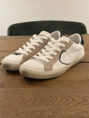 Vita sneakers  - Snygga vita sneakers med beige mockadetaljer och mörkblå häl. Skorna har vita snören, låg profil och en klassisk siluett. Materialet är en mix av läder och mocka, vilket ger en stilren och fräsch look.