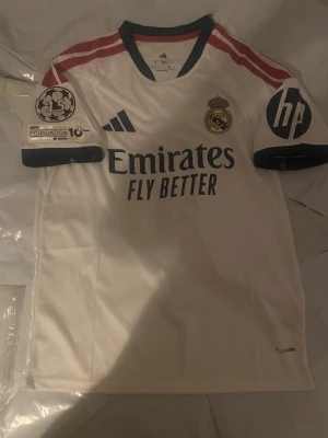 Real Madrid matchtröja Mbappé 10 - Säljer en vit Real Madrid matchtröja från Adidas med röda axelränder, klubbmärke och sponsortryck. Tröjan har trycket 'Mbappé 10' på ryggen och Champions League-märken på ärmarna. Perfekt för fotbollsfans som vill sticka ut på läktaren eller planen.