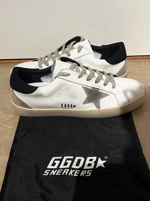 Vita GGDB sneakers med grå stjärna - Snygga vita sneakers från Golden Goose med grå stjärna på sidan, beige sula och grå skosnören. Skorna har svart detalj vid hälen och är tillverkade i läder. Perfekta för dig som gillar en clean men ändå unik look.