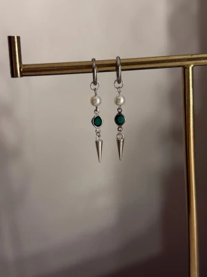 Silvriga örhängen med pärlor och gröna stenar - Säljer ett par snygga silvriga örhängen med vita pärlor, gröna runda stenar och spetsiga metallhängen. Perfekta för dig som vill ha en unik och trendig accessoar som sticker ut. Passar till många olika stilar och outfits.