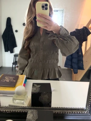Khakigrön blus med volang och puffärm - Trendig khakigrön blus med smockdetaljer över bröstet och midjan, knappar framtill och volang nedtill. Blusen har långa puffärmar och v-ringad hals. 