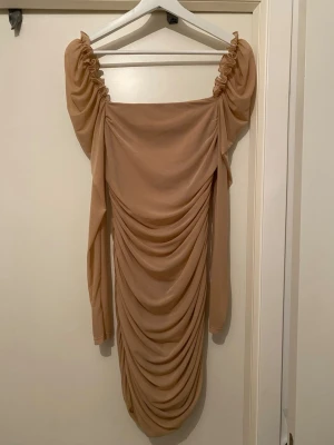 Beige långärmad draperad klänning - Snygg beige kortklänning med långa ärmar och draperad design framtill. Klänningen har rynkade axlar och en figurnära passform som framhäver siluetten. Perfekt för dig som gillar trendiga och stilrena plagg.
