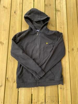 Lyle and scott hoddie - Snygg mörkgrå hoodie med dragkedja framtill och huva. Har ett litet gult broderat märke på bröstet och två fickor framtill. Perfekt för chill dagar eller när du vill ha en enkel och stilren look.