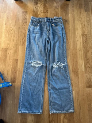 Blå raka jeans med slitningar - Säljer ett par ljusblå jeans med raka ben och stora slitningar över knäna. Jeansen har klassisk femficksmodell och hög midja. Materialet är denim i bomull och de har en avslappnad, trendig look.
