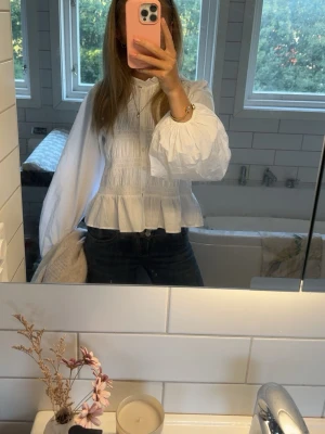 Vit blus med puffärm och smock - Säljer en vit blus med smockdetaljer och volang nedtill. Blusen har långa puffärmar och knappar framtill. Tillverkad i ett tunt, lätt tyg som ger en romantisk och trendig look. Perfekt att styla med jeans eller kjol.