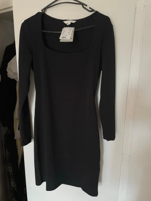 Svart långärmad klänning H&M - En stilren svart klänning från H&M med lång ärm och fyrkantig urringning. Klänningen har en enkel, figurnära passform och är tillverkad i ett mjukt material som passar perfekt till olika tillfällen.