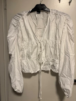 Vit blus med puffärm från Gina Tricot - Trendig vit blus från Gina Tricot i storlek XS. Blusen har puffiga långa ärmar, v-ringning och knytband framtill. Tillverkad i ett lätt bomullsliknande material med rynkade detaljer som ger en cool och avslappnad look.