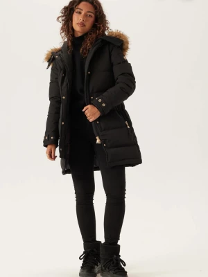 Svart lång dunjacka med fuskpäls - Snygg svart dunjacka med lång passform och huva med fluffig fuskpäls. Jackan har guldfärgade dragkedjor och knappar, två stora fickor framtill och quiltad design. Perfekt för kalla vinterdagar.