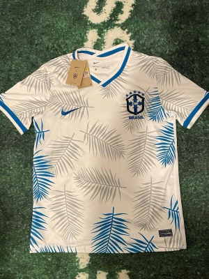 Vit Limited edition 2022 Brasilien 🇧🇷  - Snygg vit fotbollströja från Nike med blå och grå palmbladsmönster. Bröstet har Brasil-logga och Nike Swoosh i blått. V-ringad hals och blå detaljer vid ärmslut och krage. Perfekt för dig som älskar fotboll och vill sticka ut på planen. Strl S. Extra pris om du tar 2 eller fler.