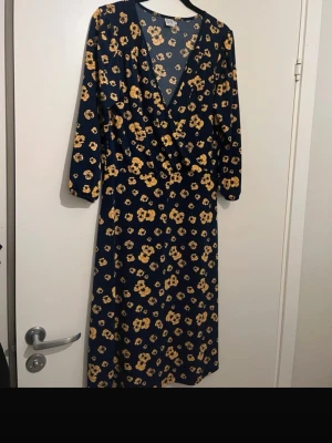 Omlottklänning - Mörkblå långklänning med gula blommor och omlottmodell. Klänningen har v-ringning och trekvartsärm, vilket ger en avslappnad men ändå stilren look. 