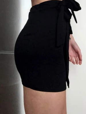 Svart skort med omlot - Helt ny, Enbart använd vid fotografering av bilderna.Stilren svart skort med hög midja och knytband i midjan. Skorten har en omlott-look framtill och en dekorativ ficka. Perfekt för dig som gillar en enkel men trendig stil. 