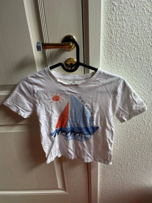 Vit t-shirt med segelbåt,122/128 - Säljer en vit croppad t-shirt med tryck av en segelbåt i blått och rött samt en liten sol. T-shirten har korta ärmar och rund hals. Perfekt för sommarens alla äventyr och ger en chill vibe.