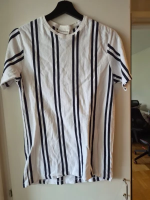 Vit randig t-shirt från Lindbergh - Säljer en fräsch vit t-shirt från Lindbergh med vertikala marinblå ränder. Klassisk rund halsringning och korta ärmar. Perfekt till jeans eller shorts för en clean och stilren look.
