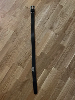 Svart Harley Davidson bälte med silverfärgat spänne, 100cm - Snyggt svart bälte i skinn med ett klassiskt silverfärgat spänne. Bältet har en stilren design och passar perfekt till jeans eller byxor. Detalj med präglad logga vid hålen ger en extra touch.