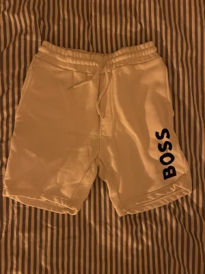 Vita shorts med BOSS-logga - Snygga vita shorts med svart BOSS-tryck på ena benet. De har elastisk midja med snörning och är gjorda i mjukt bomullsmaterial. Perfekta för chill dagar eller när du vill ha en clean look. Bakficka och avslappnad passform.