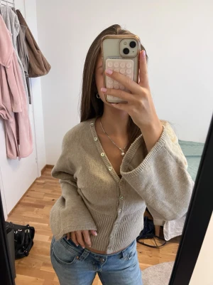 Beige ribbad kofta med v-ringning - Mysig beige kofta med v-ringning och pärlemorknappar. Koftan har långa, vida ärmar och ribbad midja som ger en snygg passform. Perfekt att matcha med jeans för en avslappnad look.