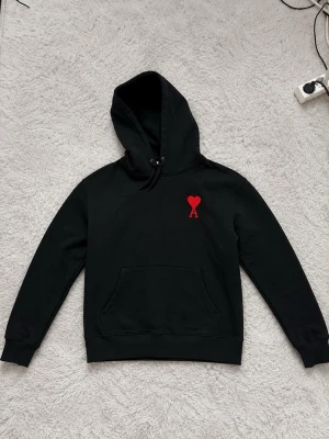 Svart ami heart hoodie  - Säljer en svart ami heart hoodie med huva och känguruficka. På bröstet finns ett broderat rött hjärta med bokstaven A under, vilket ger en unik och trendig look. Hoodien har dragsko i huvan och ribbade muddar vid ärmar och nederkant. Perfekt för dig som gillar stilrena detaljer.