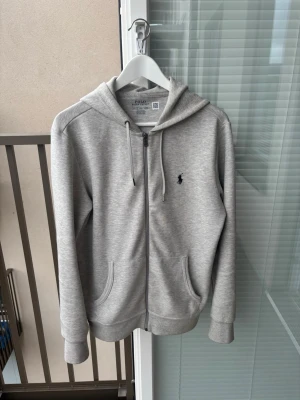 Grå tracksuit från Polo Ralph Lauren - Snygg gråmelerad hoodie med dragkedja från Polo Ralph Lauren. Klassisk design med huva och dragsnören, två fickor framtill och den ikoniska lilla loggan broderad på bröstet. Perfekt för en chill och stilren look.