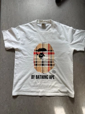 Vit BAPE t-shirt med rutig logga - Säljer en vit t-shirt från BAPE med det klassiska apansiktet i rutigt mönster på bröstet och texten 'BY BATHING APE' under. T-shirten har rund hals och är kortärmad. Perfekt för dig som gillar streetwear.