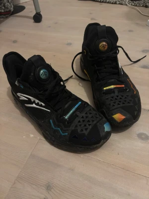 ANTA Shock Wave 5 - Unika svarta sneakers med coola geometriska mönster och färgglada detaljer i blått, orange och grönt. Skorna har hög design och snörning, samt ventilerande meshpartier och syntetiska förstärkningar. Perfekta för dig som vill sticka ut med din stil.