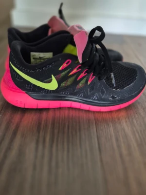 Nike svart/neon sneakers - Snygga sneakers från Nike i svart mesh med neonrosa sula och limegrönt swoosh-märke. Skorna har svarta skosnören och detaljer i rosa och grönt vid snörningen. Lätta och flexibla med sportig look, perfekta för dig som gillar färg och streetstyle.