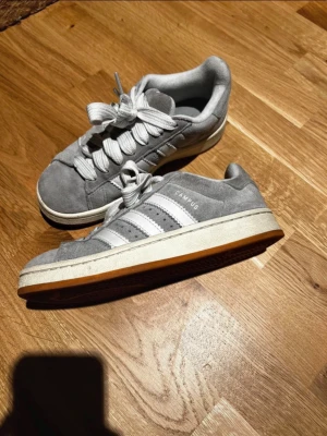 Grå Adidas Campus sneakers - Säljer ett par grå Adidas Campus sneakers i mocka med vita klassiska stripes på sidorna och tjocka vita skosnören. Skorna har låg profil och vit sula med gummidetaljer. Perfekta för en clean och stilren look.