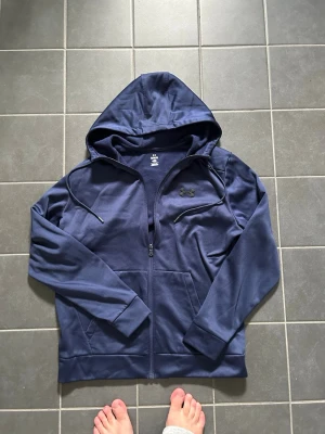 Marinblå hoodie med dragkedja - Snygg marinblå hoodie från Under Armour med dragkedja framtill, två fickor och justerbar huva med snören. Tillverkad i mjukt funktionsmaterial som passar perfekt till träning eller chill. Diskret logga på bröstet och ribbade muddar vid ärmar och nederkant.
