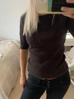 Mörkbrun ribbad topp med halvlång ärm - Säljer en mörkbrun ribbad topp med rund halsringning och halvlånga ärmar. Toppen har en tight passform och är gjord i ett mjukt, stretchigt material som sitter snyggt på kroppen. Perfekt att styla med jeans eller kjol för en clean look. Fint broderi på ärmarna. knappt använd❣️